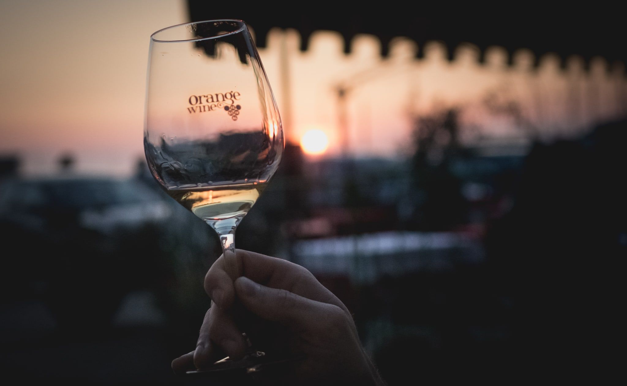 A hand holds a glass of orange wine up against the setting sun